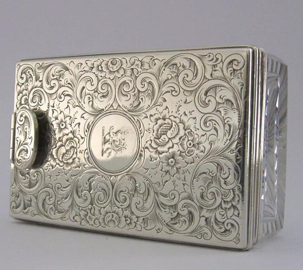 An ornate Victorian sterling silver travelling inkwell featuring floral scroll engraving and a central family crest, with a visible cut glass body.
