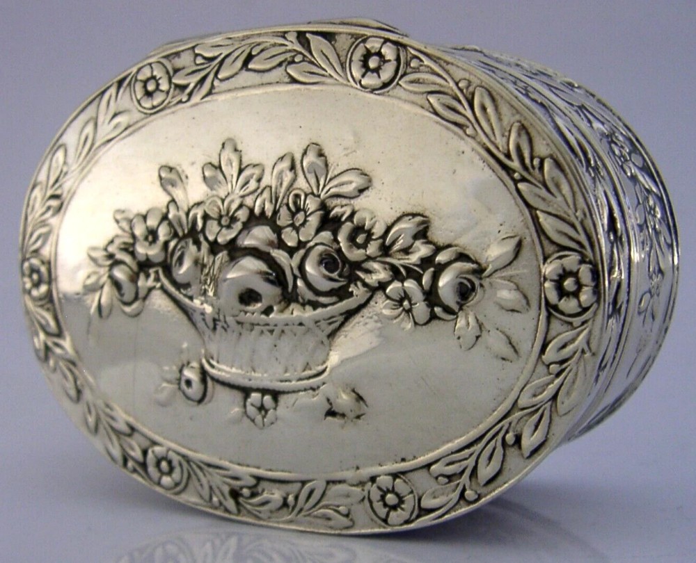 An oval solid silver table box featuring embossed flower basket motifs on the lid surrounded by a foliage border.