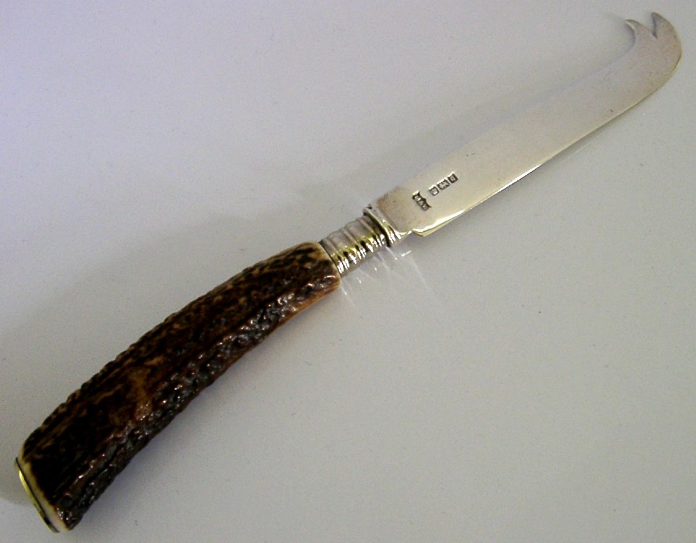 An Edwardian sterling silver cheese knife featuring a curved silver blade with hallmarks and a dark brown textured stag antler handle.