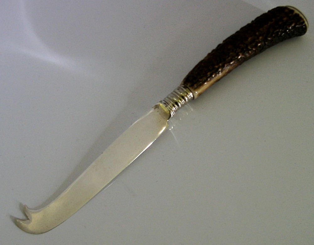 An Edwardian sterling silver cheese knife featuring a curved silver blade with hallmarks and a dark brown textured stag antler handle. View II