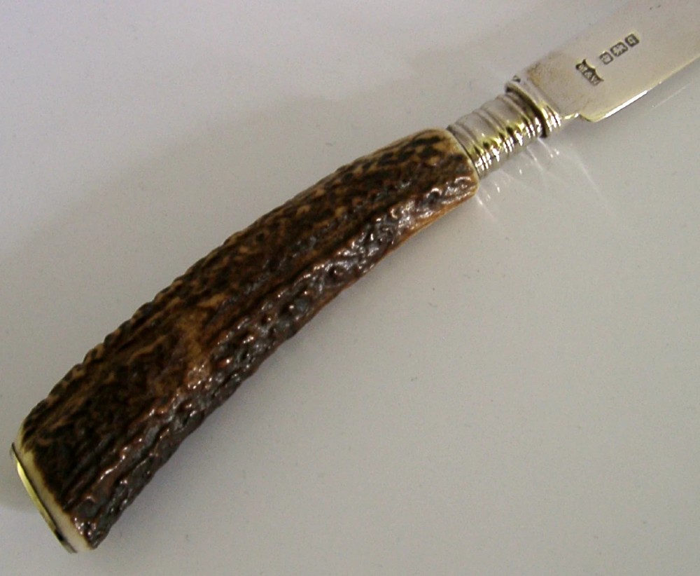 An Edwardian sterling silver cheese knife featuring a curved silver blade with hallmarks and a dark brown textured stag antler handle. View IV