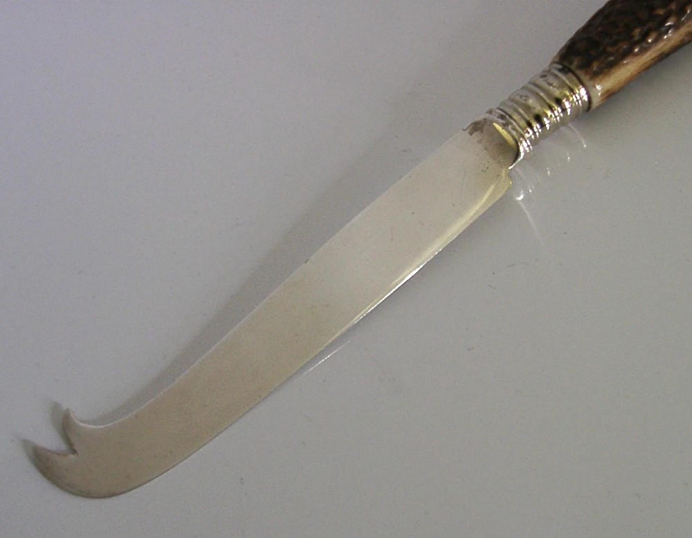 An Edwardian sterling silver cheese knife featuring a curved silver blade with hallmarks and a dark brown textured stag antler handle. View V