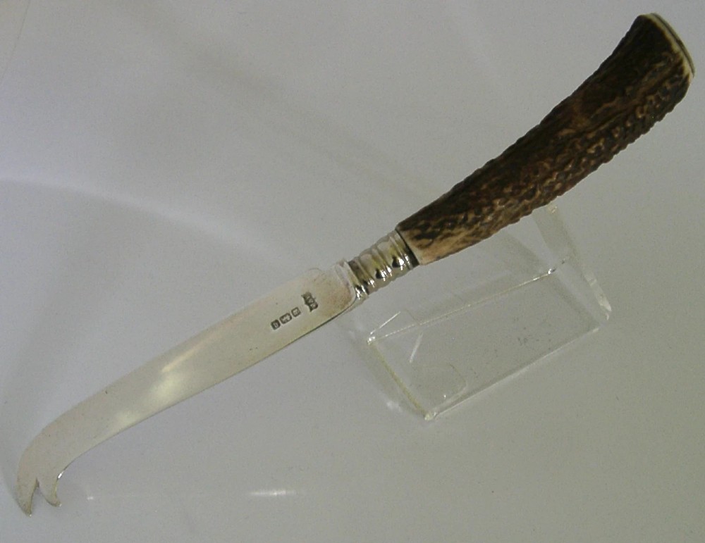 An Edwardian sterling silver cheese knife featuring a curved silver blade with hallmarks and a dark brown textured stag antler handle. View VI