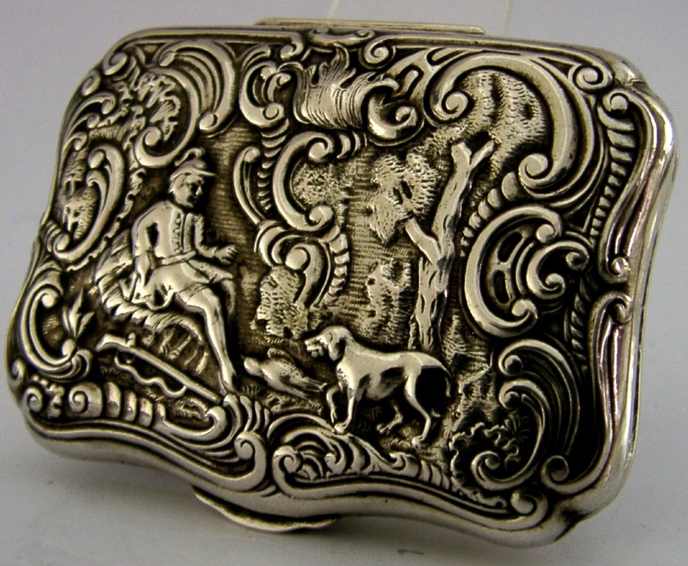 An antique rectangular silver box with intricate repousse scrollwork depicting a hunter seated beside a hound in a woodland setting.