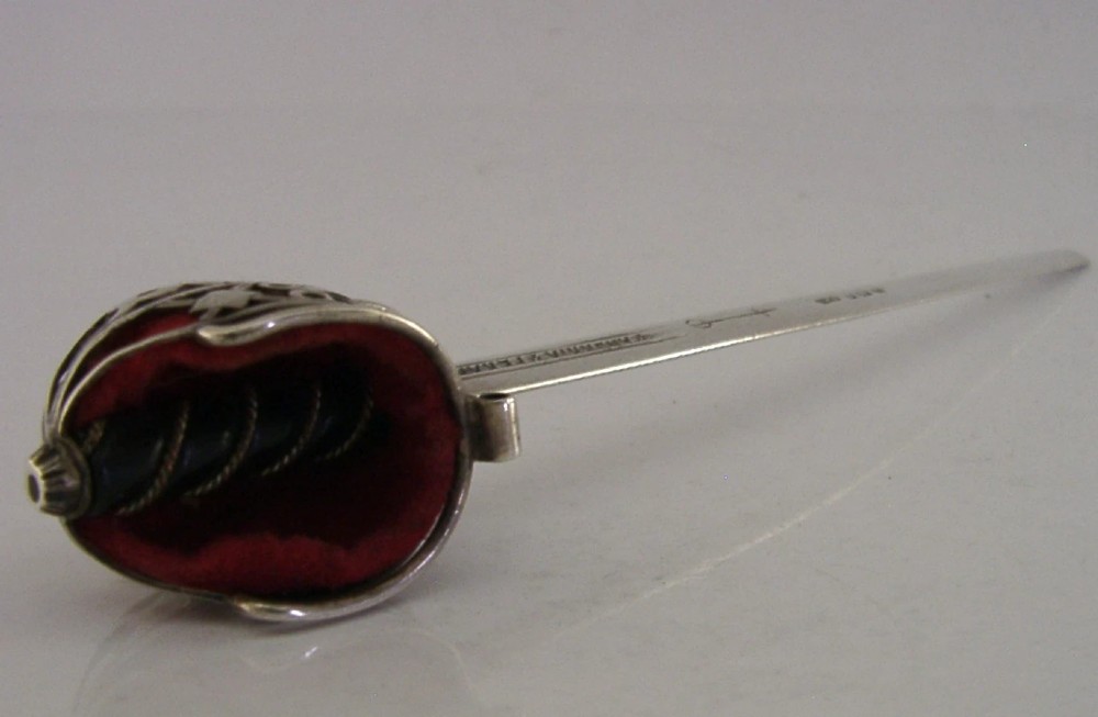 An Edwardian sterling silver letter opener featuring a smooth tapered blade stamped with British hallmarks and a decorative pierced finial housing a red orb. View X