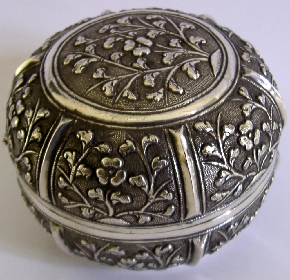 A circular antique solid silver export box decorated with hand-chased floral motifs and a stippled textured background. View X