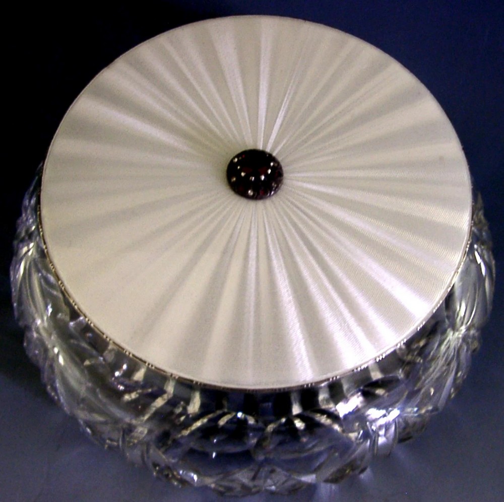 A circular cut glass dressing table jar topped with a white guilloche enamel and sterling silver lid.