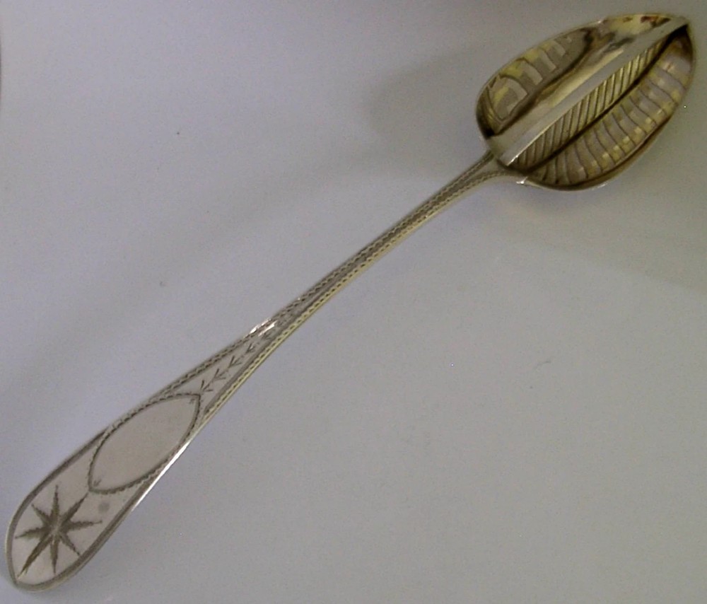 A Georgian sterling silver basting spoon with a Celtic point handle featuring bright-cut engraving and a slatted strainer divider inside the bowl.
