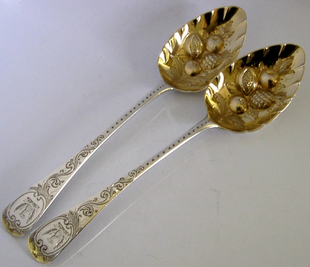 A pair of 1815 Georgian sterling silver berry spoons featuring scroll-engraved handles with family crests and gilt bowls embossed with fruit motifs.