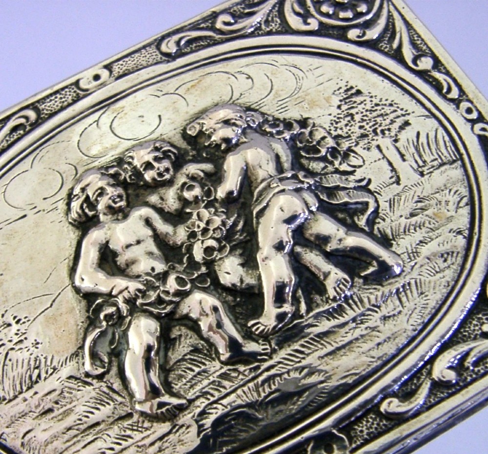 A rectangular silver trinket box featuring an embossed relief scene of three cherubs inside an oval medallion on the hinged lid. View III