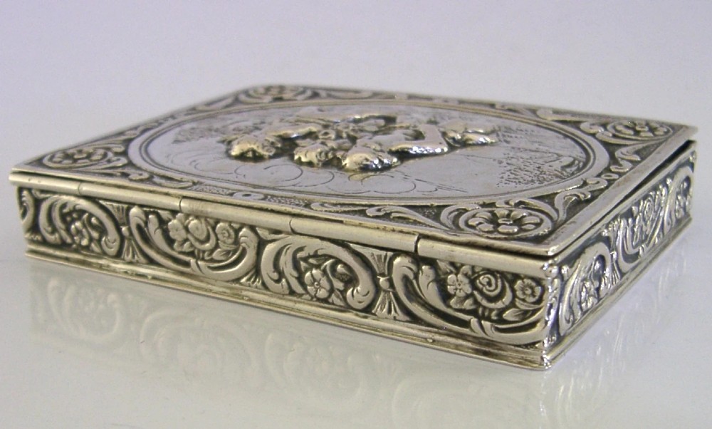 A rectangular silver trinket box featuring an embossed relief scene of three cherubs inside an oval medallion on the hinged lid. View IX
