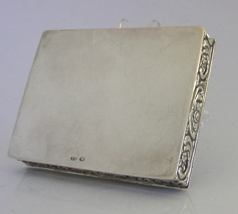 A rectangular silver trinket box featuring an embossed relief scene of three cherubs inside an oval medallion on the hinged lid. View X