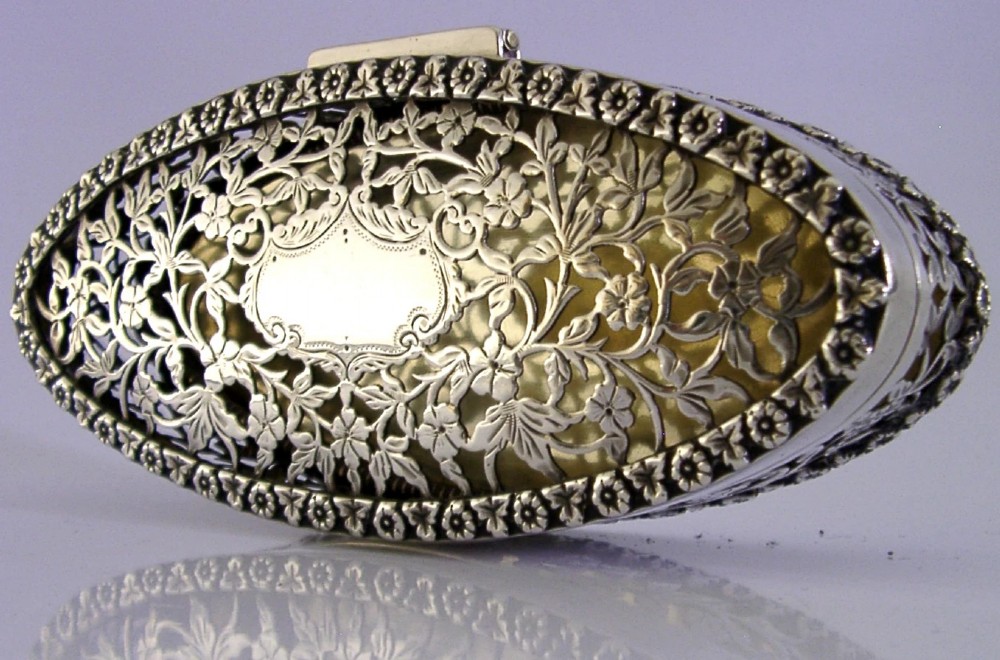 An oval sterling silver box featuring intricate pierced floral decoration, a central blank cartouche, and a gilded interior.