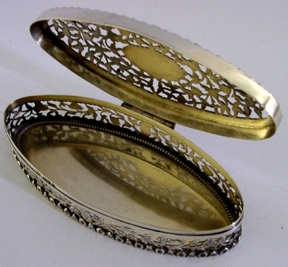 An oval sterling silver box featuring intricate pierced floral decoration, a central blank cartouche, and a gilded interior. View III