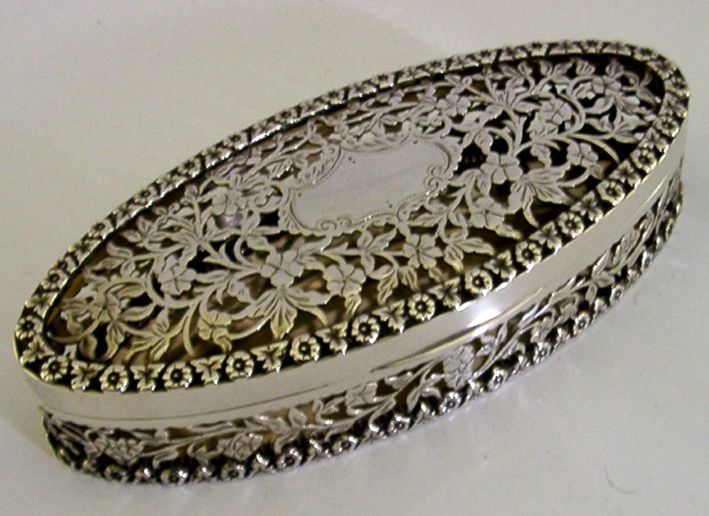 An oval sterling silver box featuring intricate pierced floral decoration, a central blank cartouche, and a gilded interior. View IV