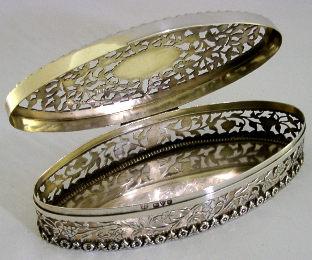 An oval sterling silver box featuring intricate pierced floral decoration, a central blank cartouche, and a gilded interior. View V
