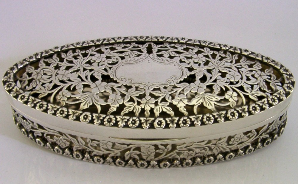 An oval sterling silver box featuring intricate pierced floral decoration, a central blank cartouche, and a gilded interior. View VI