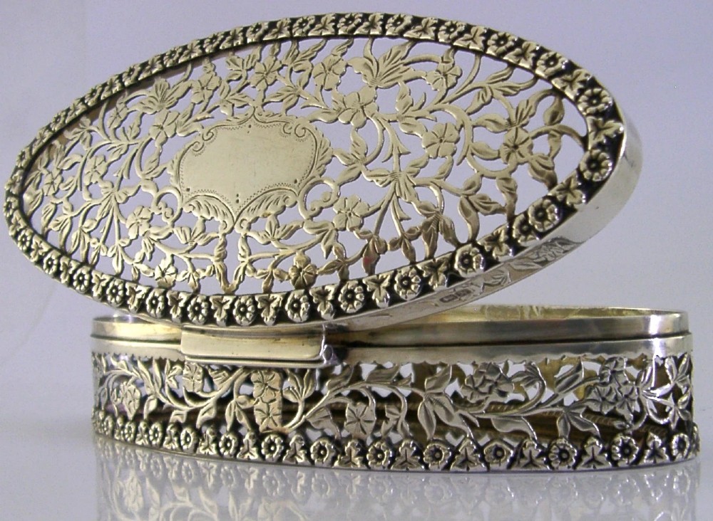 An oval sterling silver box featuring intricate pierced floral decoration, a central blank cartouche, and a gilded interior. View VII