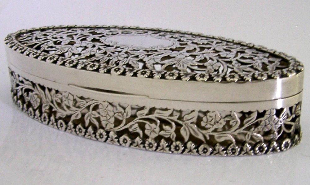 An oval sterling silver box featuring intricate pierced floral decoration, a central blank cartouche, and a gilded interior. View X