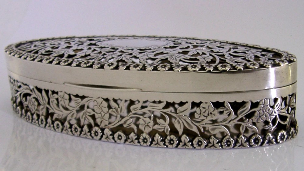 An oval sterling silver box featuring intricate pierced floral decoration, a central blank cartouche, and a gilded interior. View XII