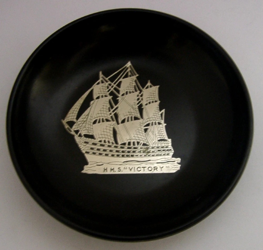 A circular black Bakelite dish featuring a central sterling silver overlay depicting the sailing ship HMS Victory.