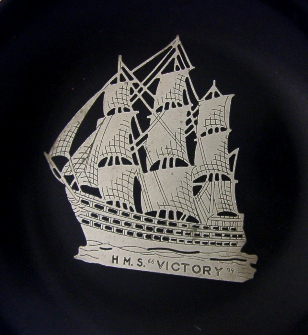 A circular black Bakelite dish featuring a central sterling silver overlay depicting the sailing ship HMS Victory. View II