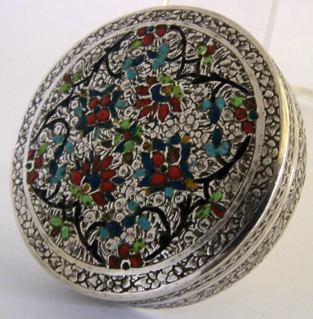 A circular solid silver trinket box with a hinged lid decorated with chased metalwork and polychrome floral enamelling in red, green, and blue.