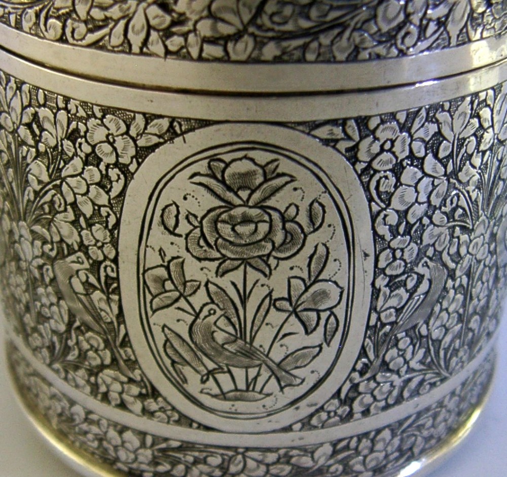 A cylindrical solid silver tea caddy featuring intricate chased floral and bird designs with a pierced pattern top lid. View V