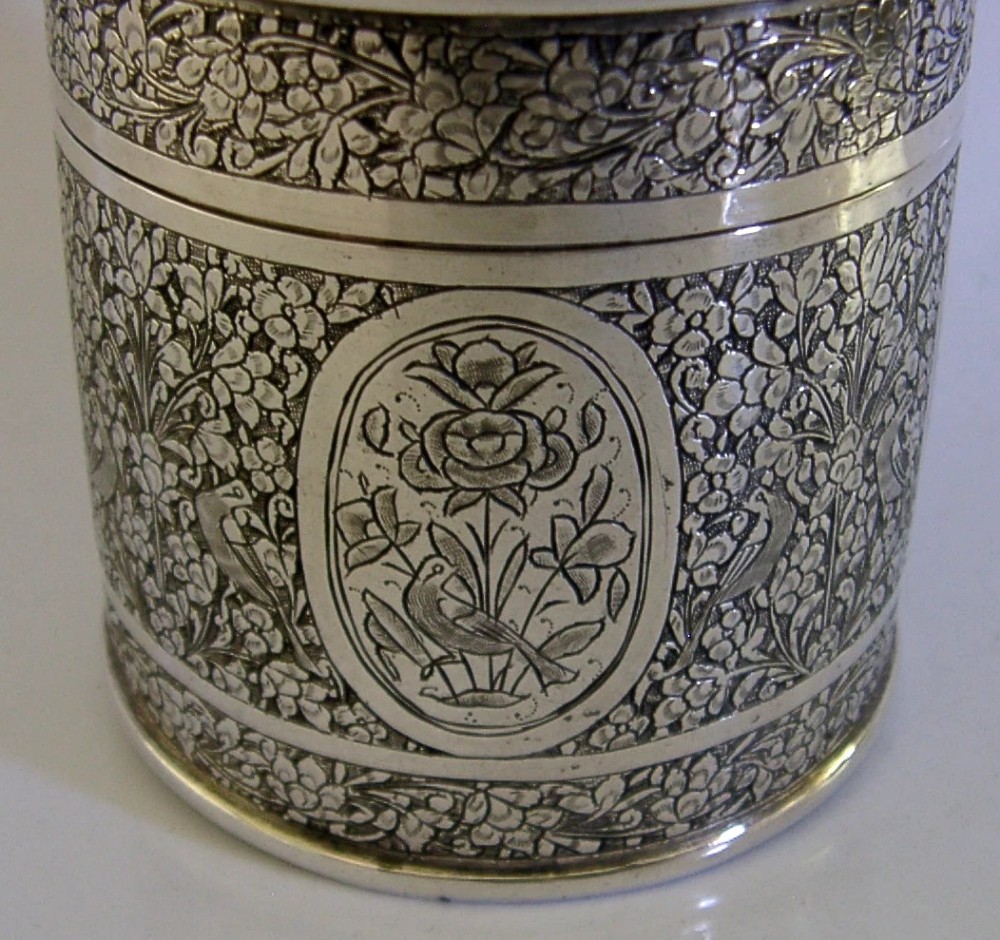 A cylindrical solid silver tea caddy featuring intricate chased floral and bird designs with a pierced pattern top lid. View VI