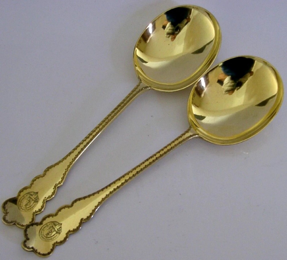 A pair of Edwardian sterling silver gilt serving spoons featuring deep oval bowls and engraved family crests on the terminals.