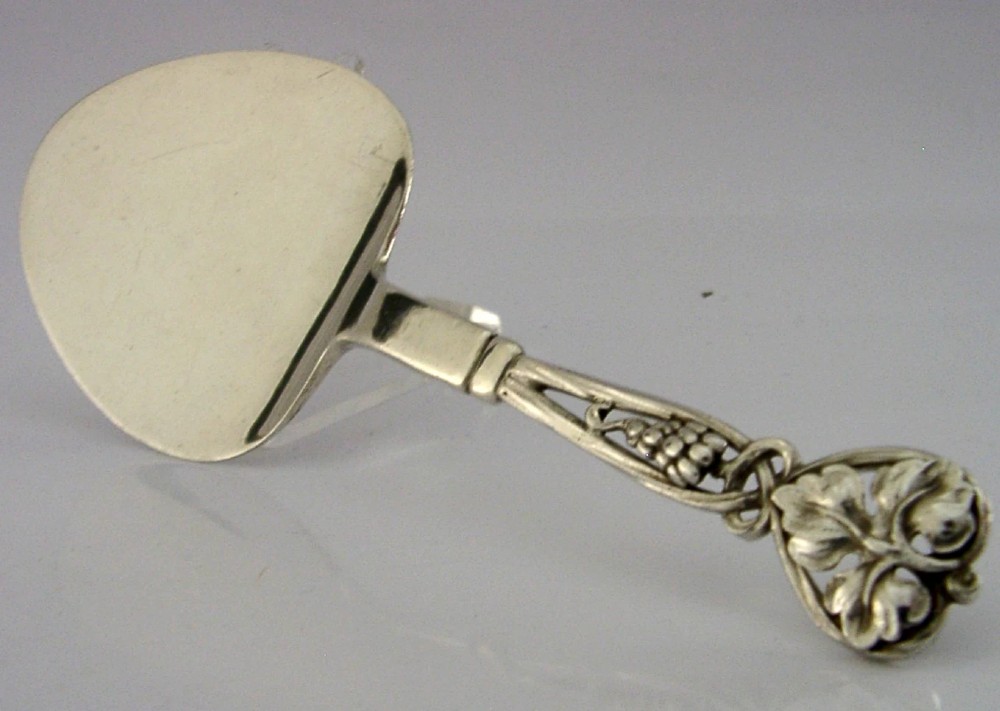 A sterling silver pastry server with a rounded flat spade blade and an openwork handle decorated with carved leaf and grape cluster motifs.