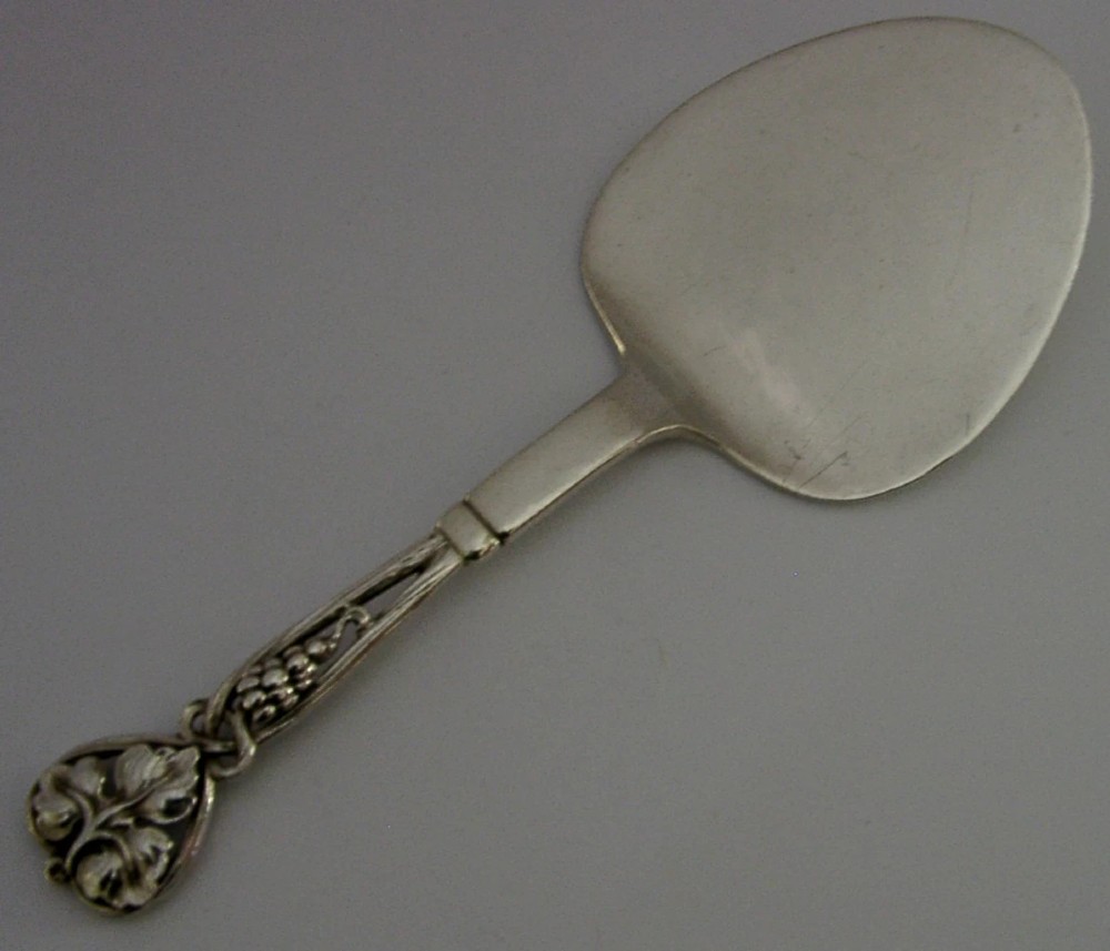 A sterling silver pastry server with a rounded flat spade blade and an openwork handle decorated with carved leaf and grape cluster motifs. View IV