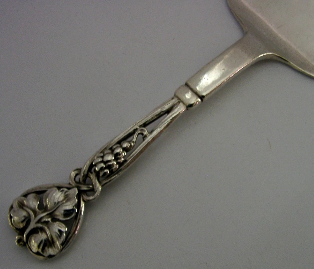 A sterling silver pastry server with a rounded flat spade blade and an openwork handle decorated with carved leaf and grape cluster motifs. View V