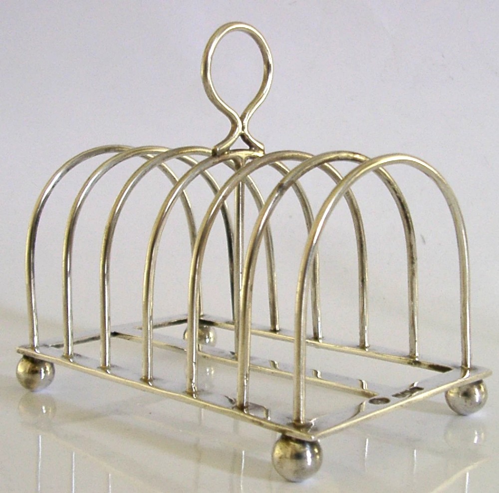 A sterling silver six-slice toast rack featuring seven arched wire divisions, a central loop handle, and four ball feet resting on a rectangular base.