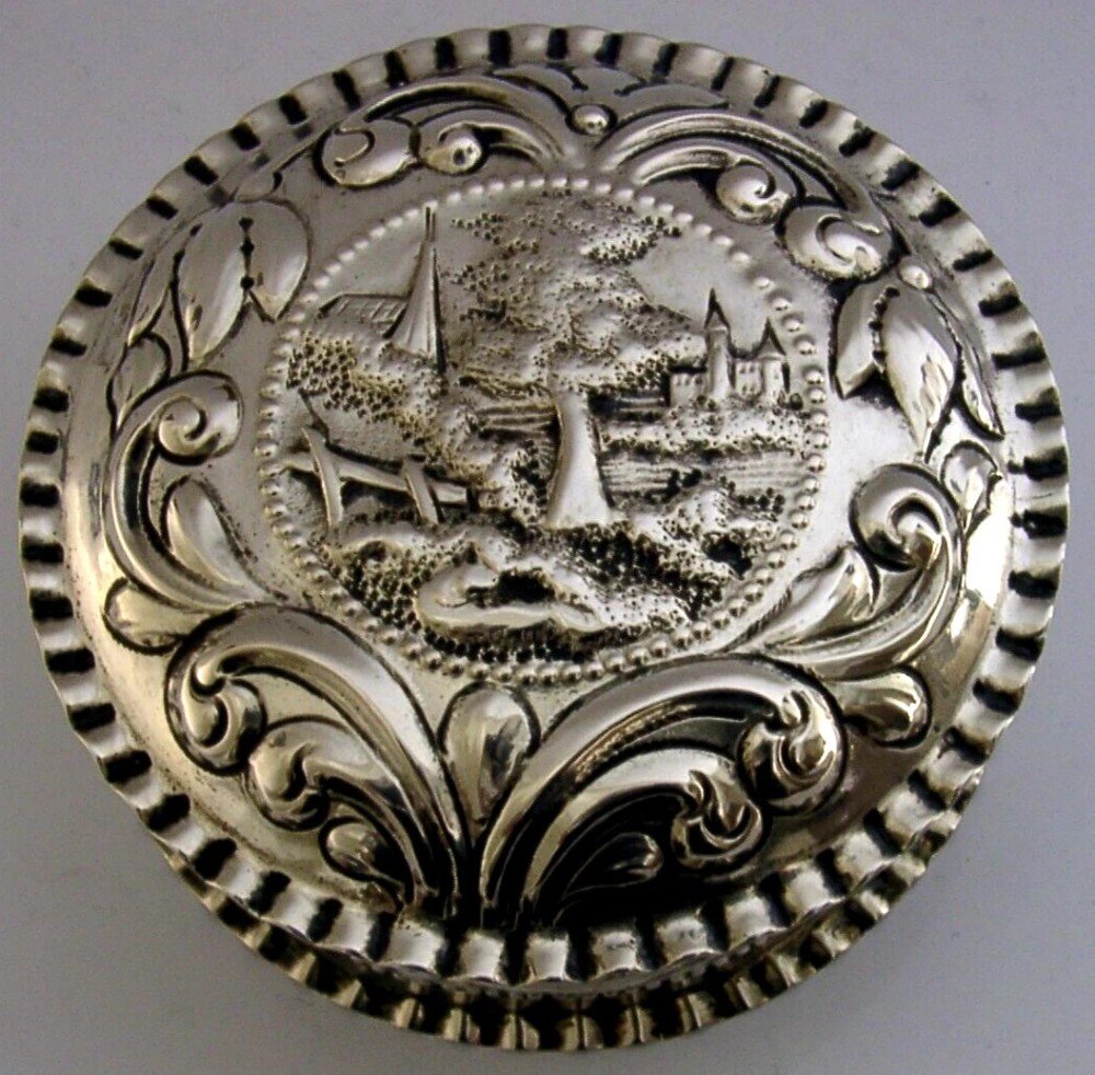 A circular solid silver table box featuring embossed repousse detail of a castle scene framed by acanthus scrolls and fluted edges.