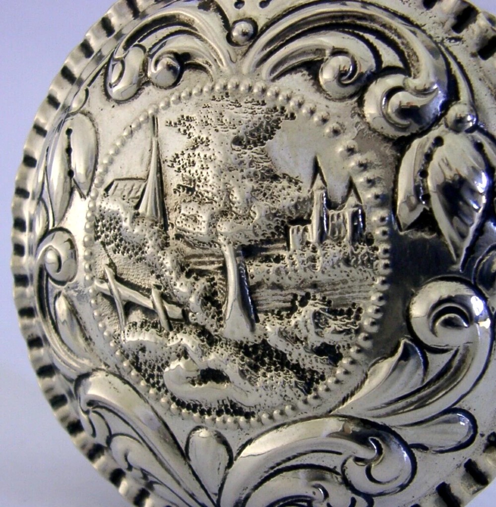 A circular solid silver table box featuring embossed repousse detail of a castle scene framed by acanthus scrolls and fluted edges. View II