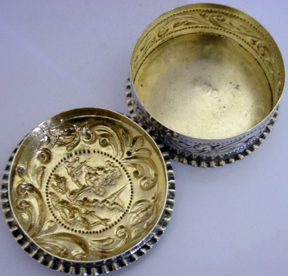 A circular solid silver table box featuring embossed repousse detail of a castle scene framed by acanthus scrolls and fluted edges. View III