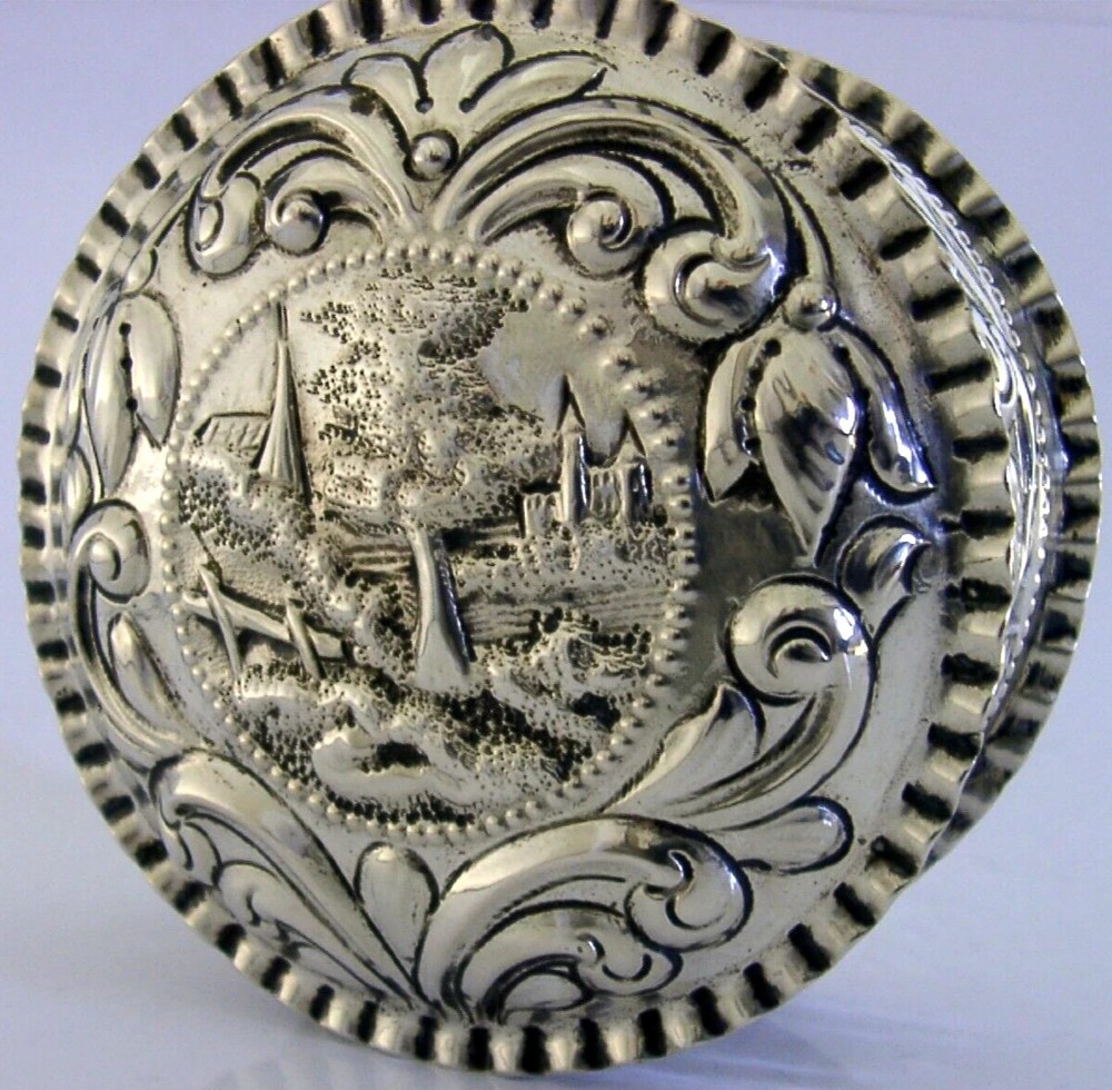 A circular solid silver table box featuring embossed repousse detail of a castle scene framed by acanthus scrolls and fluted edges. View IV