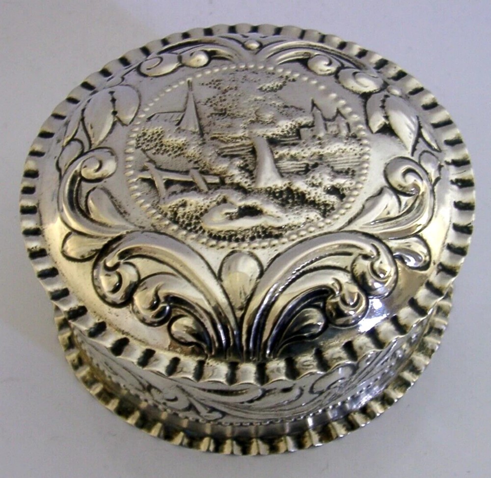 A circular solid silver table box featuring embossed repousse detail of a castle scene framed by acanthus scrolls and fluted edges. View V