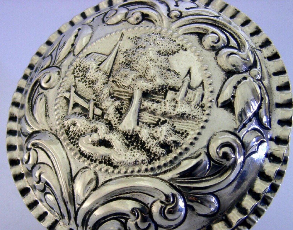 A circular solid silver table box featuring embossed repousse detail of a castle scene framed by acanthus scrolls and fluted edges. View VI