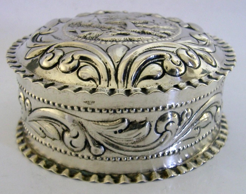 A circular solid silver table box featuring embossed repousse detail of a castle scene framed by acanthus scrolls and fluted edges. View VII
