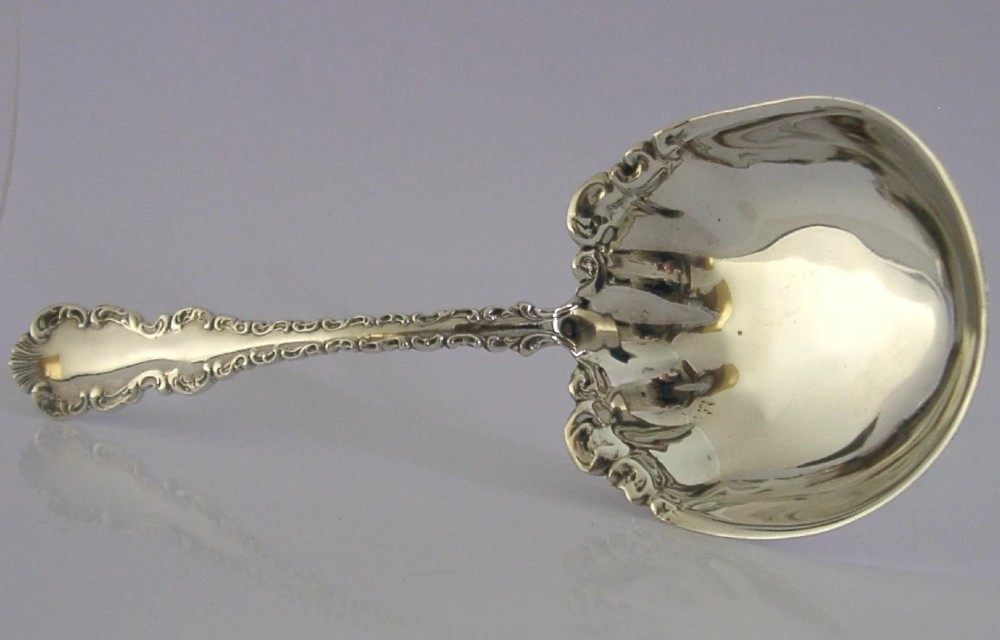 An antique sterling silver serving spoon featuring a scalloped shell-shaped bowl and an ornate scrollwork handle.