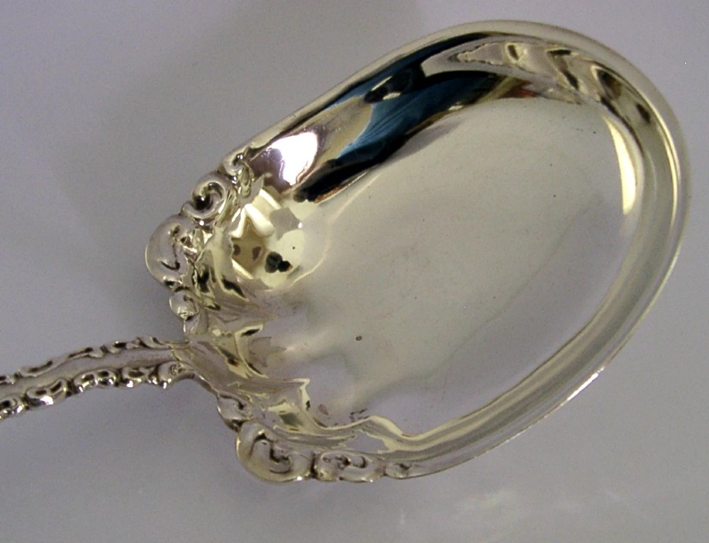 An antique sterling silver serving spoon featuring a scalloped shell-shaped bowl and an ornate scrollwork handle. View II