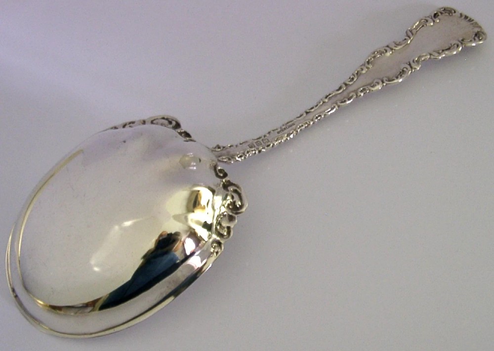 An antique sterling silver serving spoon featuring a scalloped shell-shaped bowl and an ornate scrollwork handle. View IV