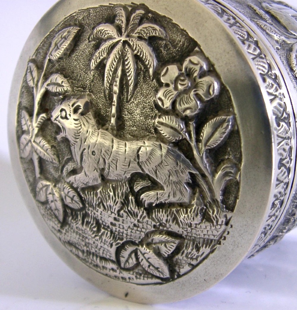 A cylindrical antique Indian solid silver caddy box with a fitted lid, decorated with embossed repousse relief depicting trees, foliage, and village huts. View IV