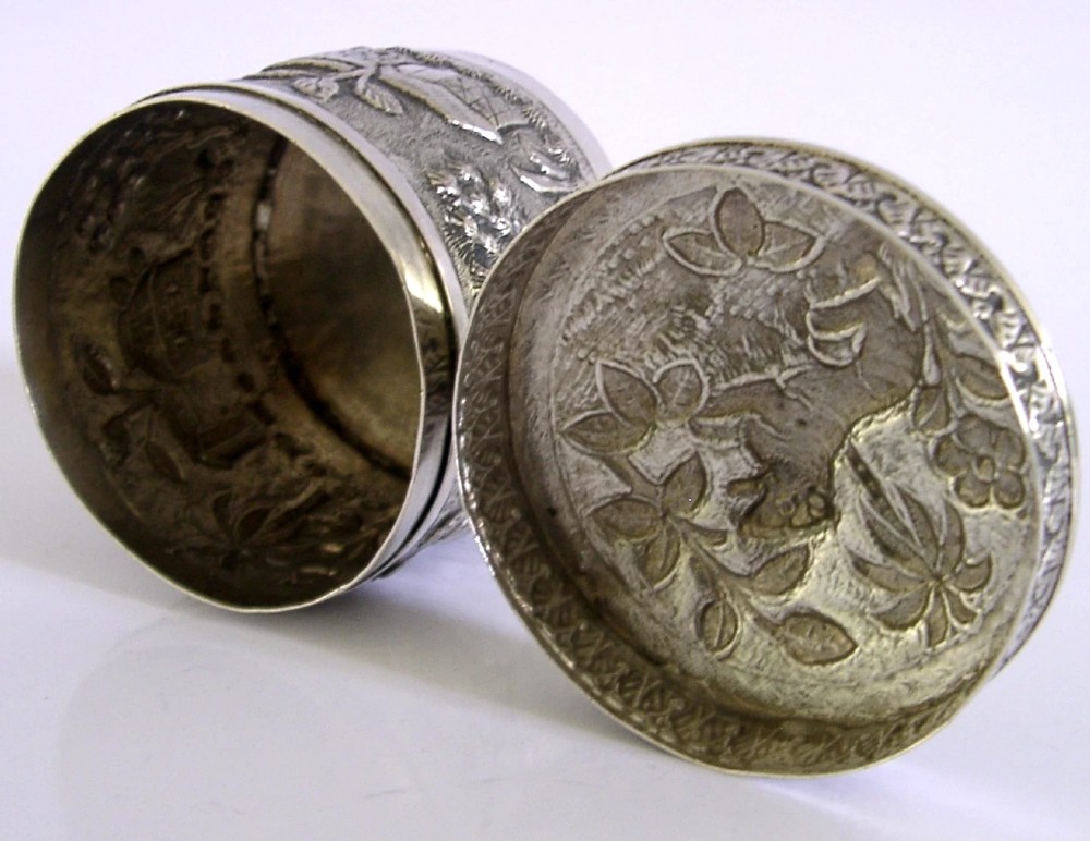 A cylindrical antique Indian solid silver caddy box with a fitted lid, decorated with embossed repousse relief depicting trees, foliage, and village huts. View V