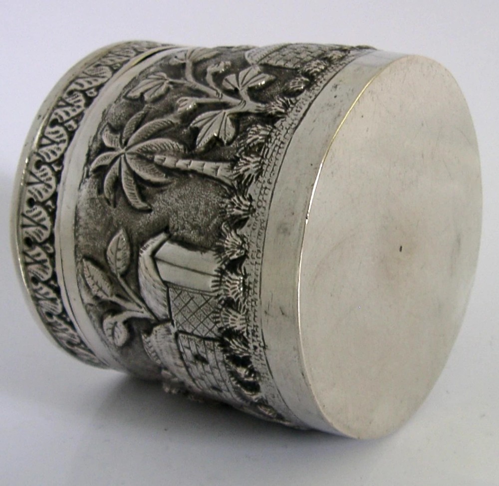 A cylindrical antique Indian solid silver caddy box with a fitted lid, decorated with embossed repousse relief depicting trees, foliage, and village huts. View VII