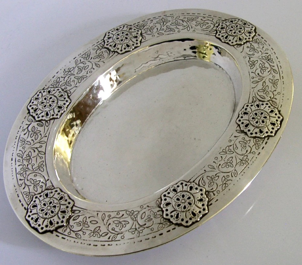 An oval antique sterling silver dish featuring a planished centre and a wide rim decorated with chased floral rosettes and leaf motifs.