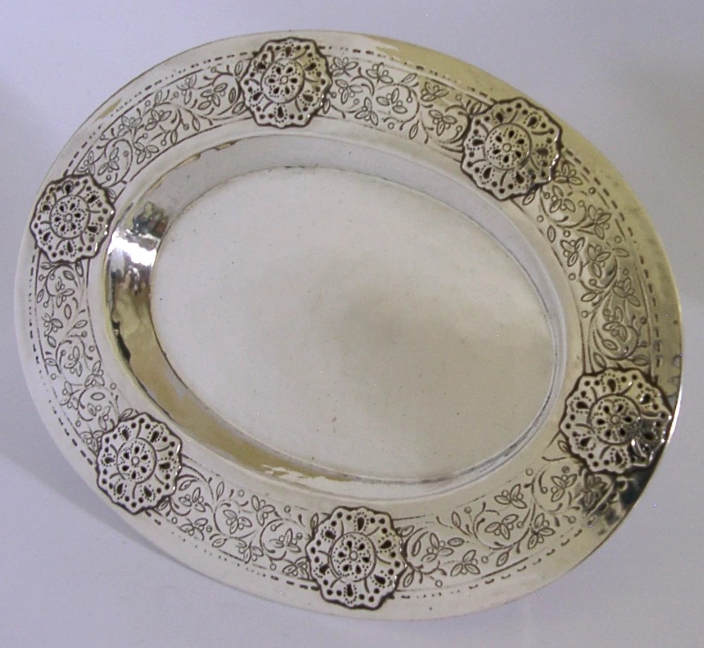 An oval antique sterling silver dish featuring a planished centre and a wide rim decorated with chased floral rosettes and leaf motifs. View IV