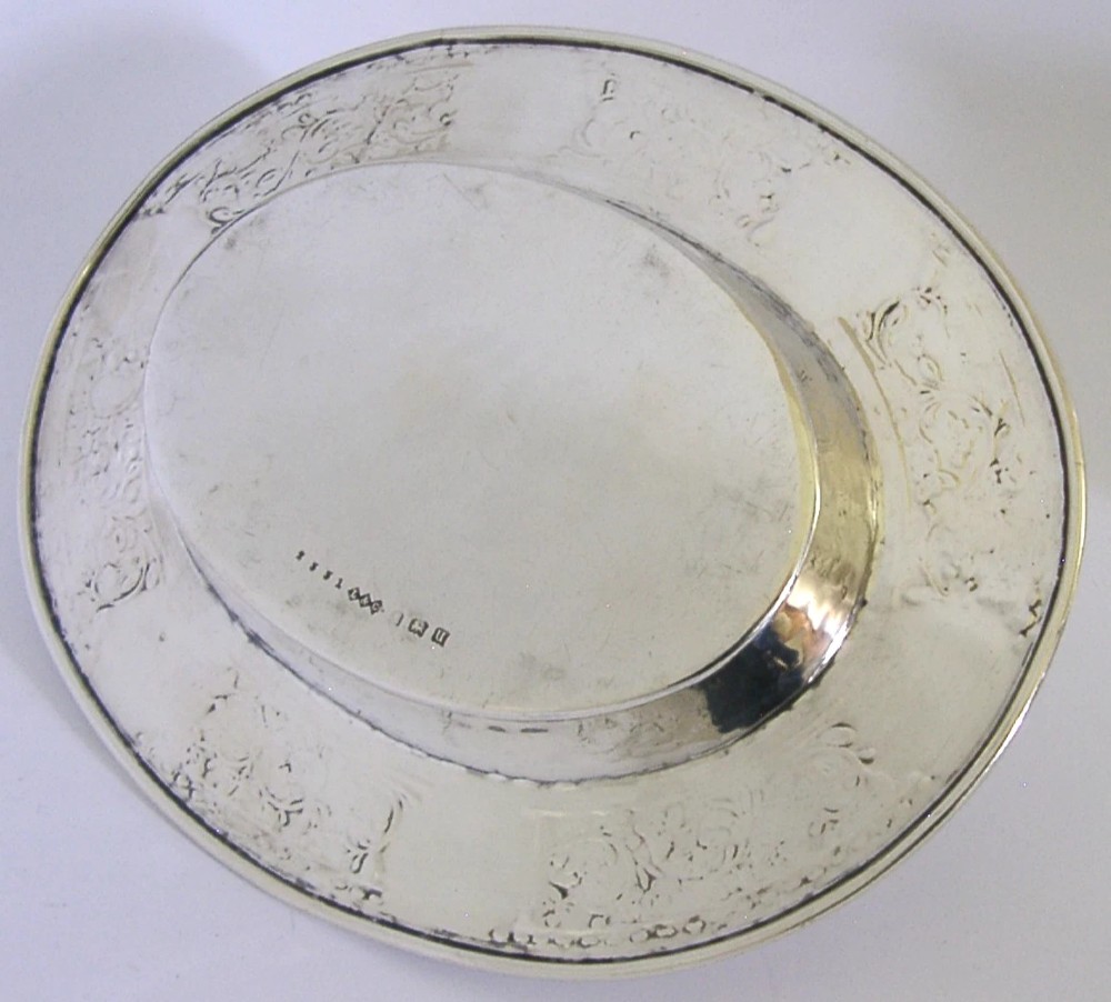 An oval antique sterling silver dish featuring a planished centre and a wide rim decorated with chased floral rosettes and leaf motifs. View V